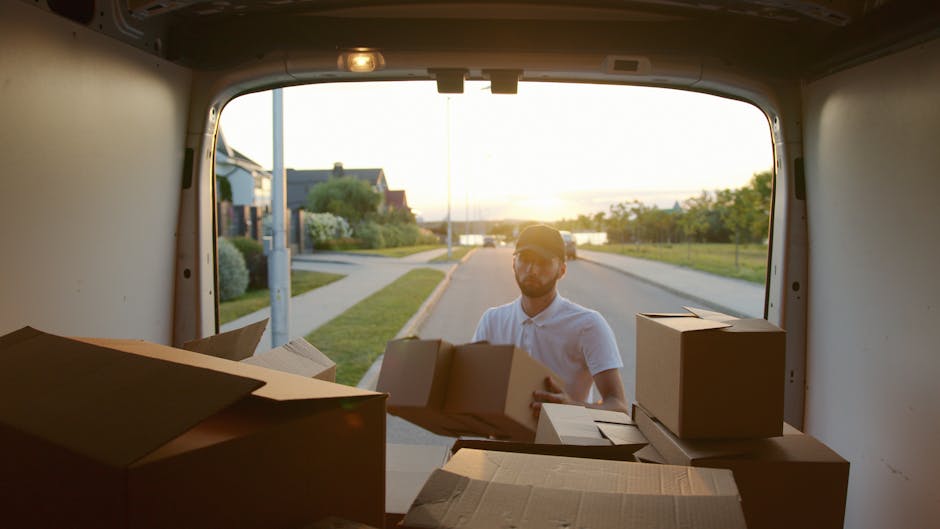 A man in a white shirt and cap is loading cardboard boxes into the back of a large white moving van during a home relocation. The van's interior is visible, with several packed boxes made of brown car