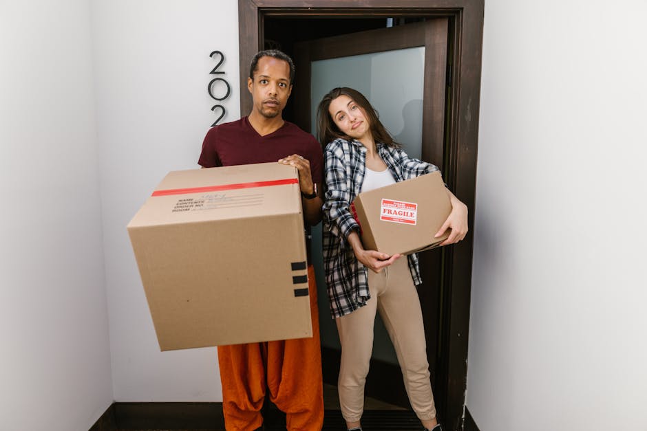 A man and a woman standing inside a doorway in a residential property, each holding a cardboard box used for home relocation. The man, positioned on the left, has a serious expression and is carrying a large box wrapped with red tape, with some black markings visible. The woman, on the right, leans her head slightly on the doorframe with a relaxed expression while holding a smaller box labeled 'FRAGILE' with red and white packaging tape. She is dressed casually in a plaid shirt and beige trousers. The background shows a dark wooden doorframe and a plain white wall, with natural lighting illuminating the scene. The image captures the loading process of packing and moving tasks, illustrating staff from Man and Van Barnes involved in furniture transport and home removals at an apartment entrance, with some cardboard packaging materials and the doorway indicating ongoing household clearance or an imminent move.