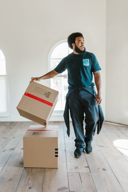 A man wearing a dark blue t-shirt with a logo on the chest and dark trousers is inside a well-lit room with light-colored walls and wooden flooring. He is holding a cardboard box wrapped with red packing tape in his right hand, preparing to place it on top of another box on the floor. The room has a large arched window allowing natural light to fill the space, indicating an empty property possibly during a home relocation process. The scene depicts the loading and packing stage of house removals, with clean, sturdy cardboard boxes suitable for moving household items. The presence of the boxes and the man's actions suggest an organized packing and furniture transport operation occurring as part of a professional removals service, such as those offered by Man and Van Barnes, specializing in urgent and same-day move assistance for areas like Barnes.