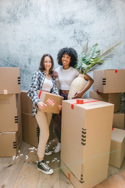 A man and a woman standing inside a doorway in a residential property, each holding a cardboard box used for home relocation. The man, positioned on the left, has a serious expression and is carrying a large box wrapped with red tape, with some black markings visible. The woman, on the right, leans her head slightly on the doorframe with a relaxed expression while holding a smaller box labeled 'FRAGILE' with red and white packaging tape. She is dressed casually in a plaid shirt and beige trousers. The background shows a dark wooden doorframe and a plain white wall, with natural lighting illuminating the scene. The image captures the loading process of packing and moving tasks, illustrating staff from Man and Van Barnes involved in furniture transport and home removals at an apartment entrance, with some cardboard packaging materials and the doorway indicating ongoing household clearance or an imminent move.